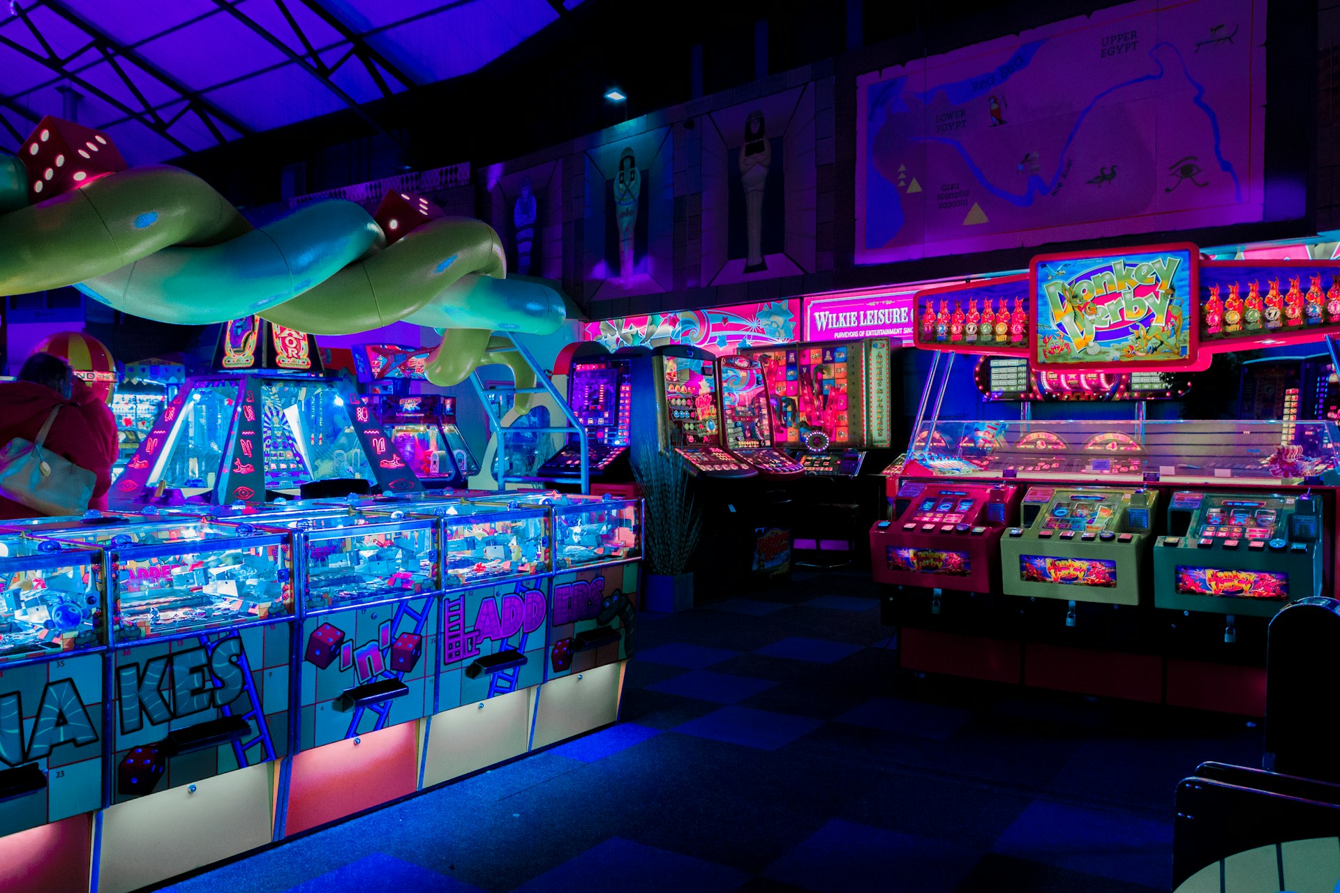An indoor arcade filled with vibrant gaming machines lit with neon blue and pink lights. The scene includes various arcade games with flashing screens and colorful displays, and a large overhead structure resembling a green snake. The background shows illuminated signs and themed decor, contributing to an immersive entertainment atmosphere.