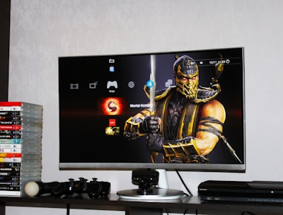 A flat-screen TV displays a video game interface featuring a character from Mortal Kombat in the center. Beside the TV is a stack of video game cases for the PlayStation 3, including titles like FIFA 14 and Grand Theft Auto. Below the TV, there is a gaming console, controllers, and other gaming equipment.