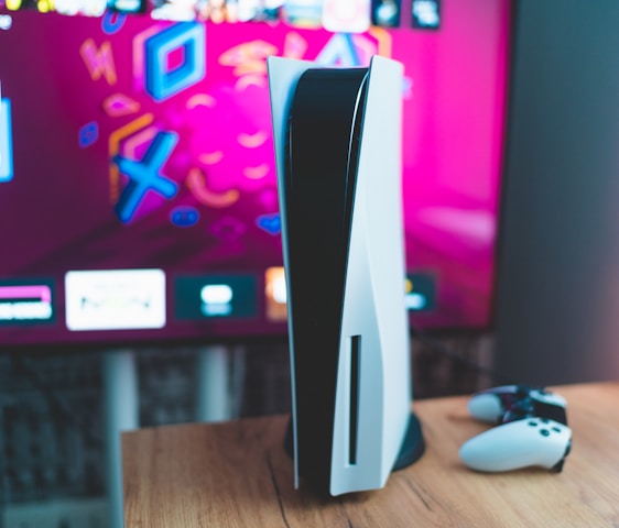 A gaming console is placed on a wooden table with a game controller nearby. In the background, a television screen displays colorful graphics, including geometric shapes and vibrant colors.